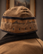 A close-up of a brown canvas beanie with black trim and embroidered text reading 'Stormy Kramer + DECOY' on a person's head, paired with a brown jacket.