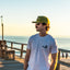Man wearing olive Pintail Rope Hat and gray DECOY t-shirt standing on a beach boardwalk at sunset
